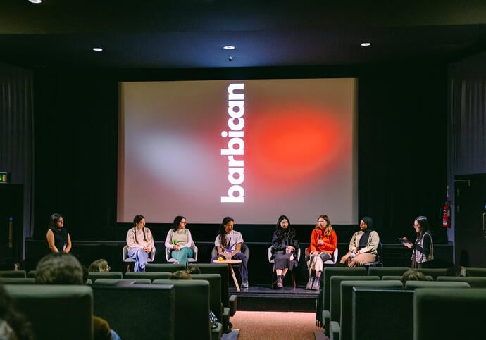 image of a panel seated in front of a cinema screen which has the Barbican logo displayed