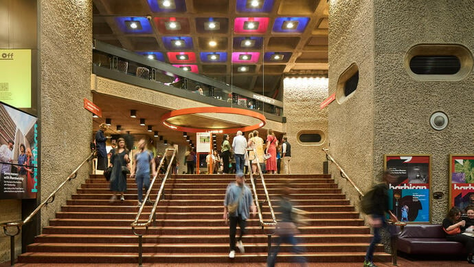 Blurred people walk down the stairs from the Barbican foyers, with bright ceiling panel lights