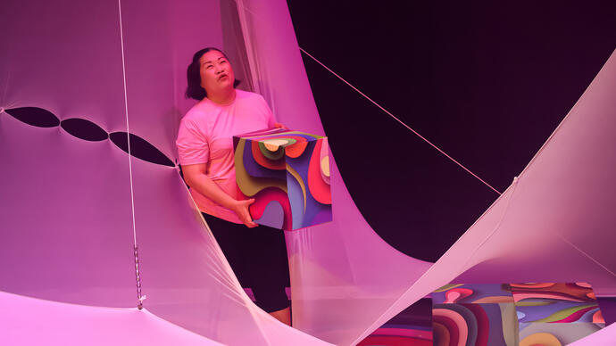 A person stands holding a square box lit with bright colours while standing among large, white draping fabric.