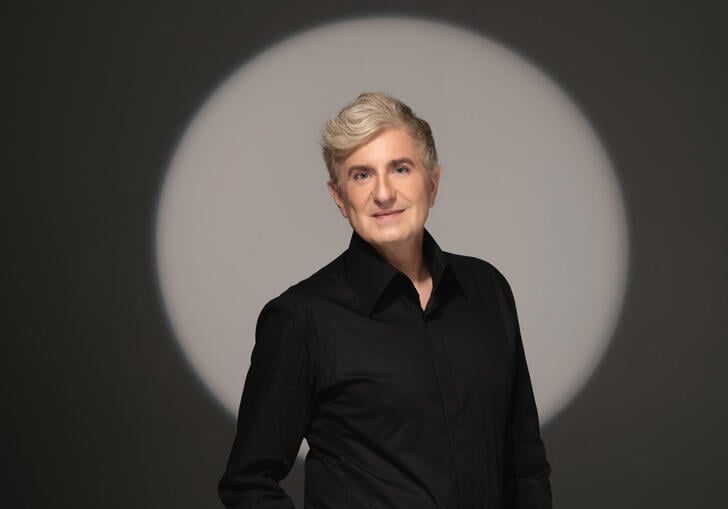 Jean Yves Thibaudet standing in front of a grey background with a spotlight forming a light circle behind him