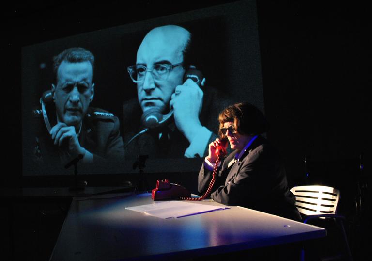 A photo showing a character called Lois Weaver in glasses, wearing a black jacket on the phone. It is a red coloured phone and is on a table with some paper. In the background there is a clip from Dr. Strangelove the film