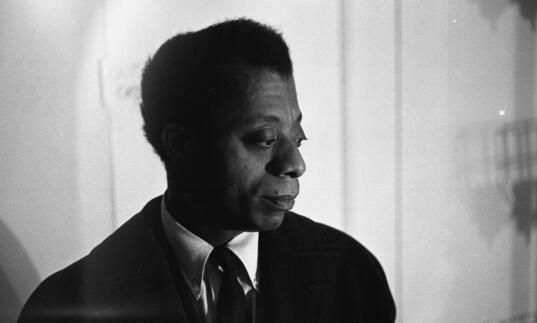 James Baldwin, wearing a suit, looks thoughtfully to the side. 