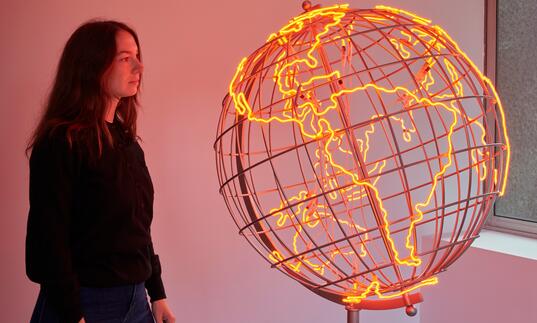 A woman looks at a sculpture of a globe, the outlines of the countries lit in neon orange strip lights.