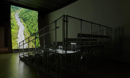 In a dark gallery, metal bleacher-style seating is positioned in front of a large, vertical, curved screen showing a river coursing through a lush, forested valley.