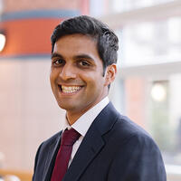 A headshot of the board member Aaron D'Souza, smiling and wearing a suit and tie