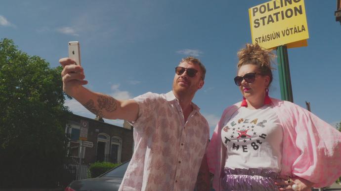 a man and a woman dressed in pink take a selfie with the polling station sign behind them