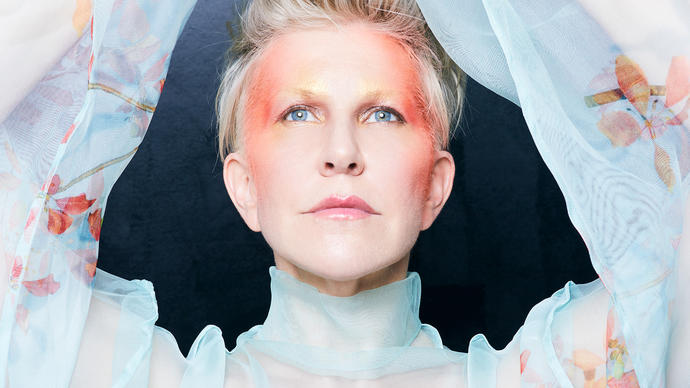 Joyce DiDonato in a blue floaty top with her arms above her head, with red face paint on her temple and the sides of her face