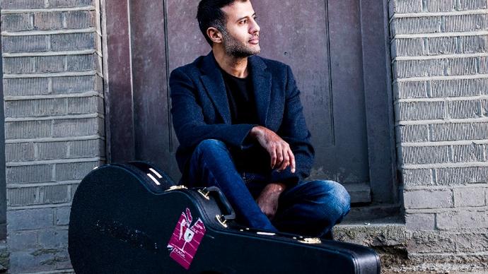 colour photo of Hamza Namira sitting on a step with his guitar case