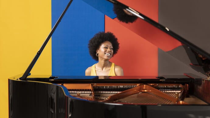 Isata Kanneh-Mason sits at a piano, smiling and looking left. Behind her is a backdrop of yellow, blue, red and black stripes. 
