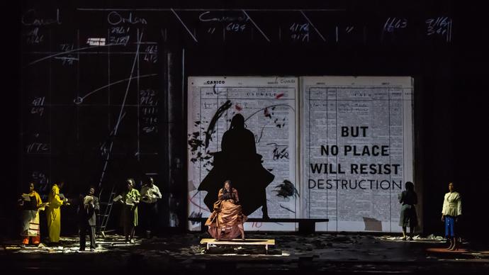 A performer in an orange dress dances in front of a large projection of a book. Other performers stand to the left-hand side and sing. 