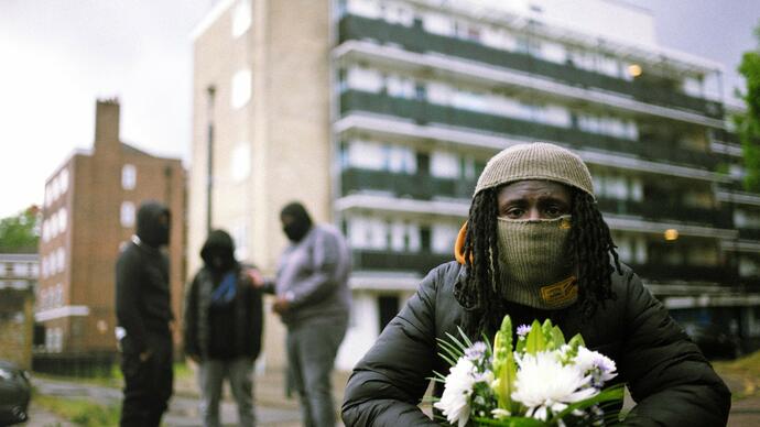 A man wearing a face covering holds white flowers in front of a block of flats. Three men stand in a group behind him. 