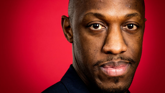 Actor Giles Terera looks straight at the camera against a red background.