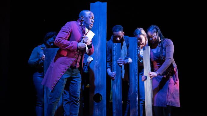 Actor Giles Terera leans against a wooden post while other performers stand with their heads down. 