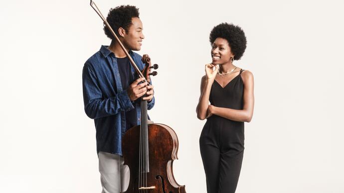 Sheku and Isata Kanneh-Mason smiling at each other. Sheku is holding his cello and bow and they are standing in front of a white background.