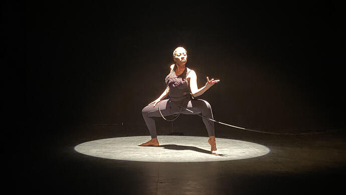 Performer Julene Robinson stands in a spotlight with her arm outstretched towards the audience. 