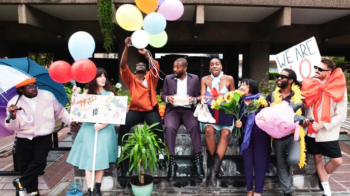 The PappyShow company sit on a wall at the Barbican Centre with signs, balloons, flowers and other objects celebrating the fact they are 10.