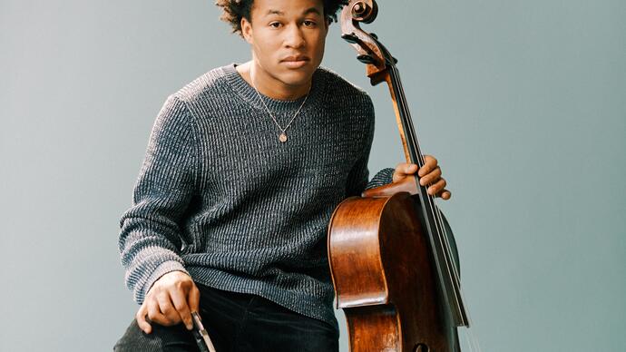 Sheku Kanneh-Mason sitting on a wooden stool with his cello, in front of a pale blueish gray background