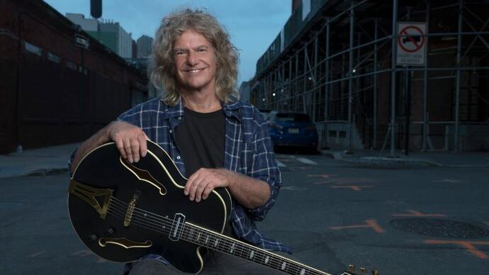 Pat Metheny sits, holding his guitar, on the street. 