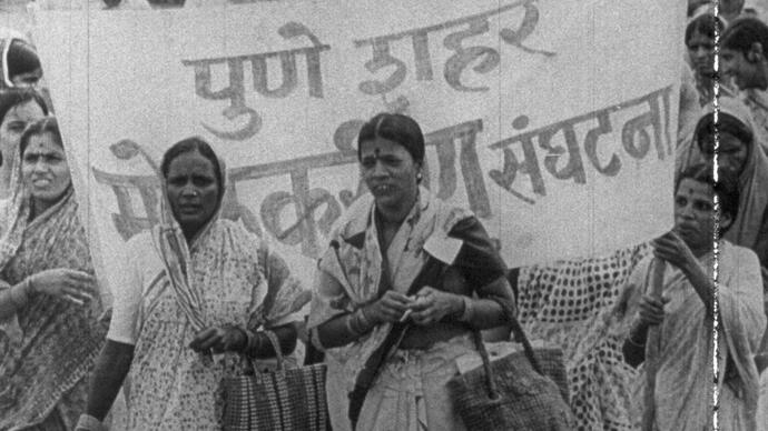 Women stand infront of a protest banner marching down the street.