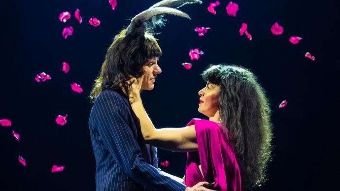 Two people embrace as pink petals fall around them. One person wears antlers on their head.