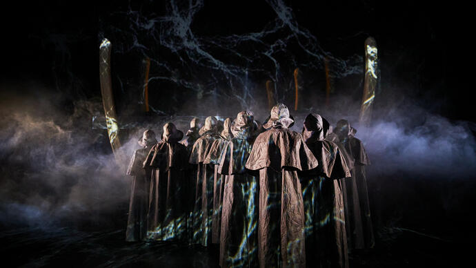 Group of people in brown cloaks facing away from the camera, smoke behind.