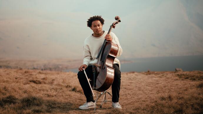 Sheku Kanneh-Mason sitting with his cello in a mountainous landscape, with a lake in the distance behind him