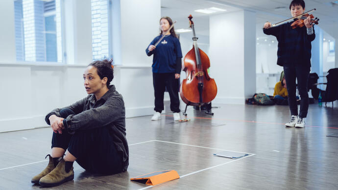 Ruth Negga sits on the floor of the rehearsal room as two people play string instruments behind them.