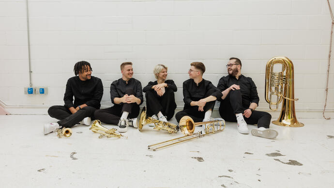 The members of Connaught Brass sitting in a row in front of a white wall, their instruments placed on the floor in front of them.