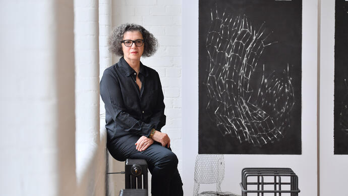 Mona Hatoum sitting on a radiator on the wall of her studio. On the wall next to her is Untitled (Bed Springs), one of her lithograph print depicting a structure made of bed springs, curved and crumpled.