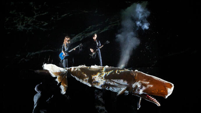 A whale-sized whale puppet floats on stage in dark water, two people play string instruments behind it,