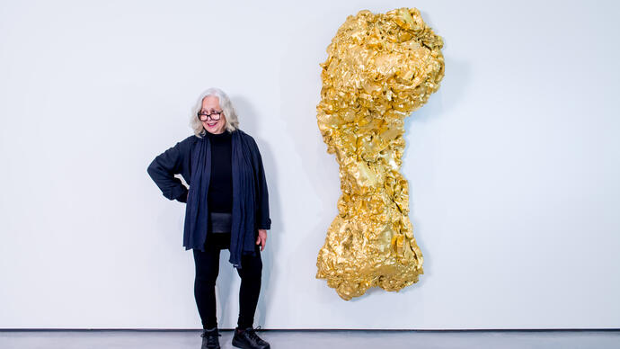 Lynda Benglis standing next to one her sculpture titled Ghost Dance/Pedmarks. It is a large, gold, torso-like form hanging on a white wall.