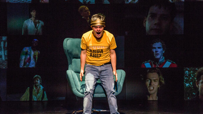 A person in a yellow shirt and blue jeans standing over a green sofa chair, shouting. They are against a background of screens with each screen showing pictures of different men.