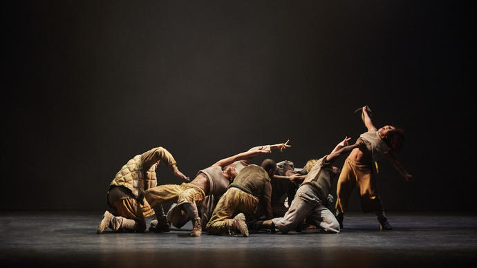 Image of a group of dancers in various positions. Most of them are crouched and reaching towards a dancer at the far right who is upright. All the dancers are in brownish and/ or grey clothing