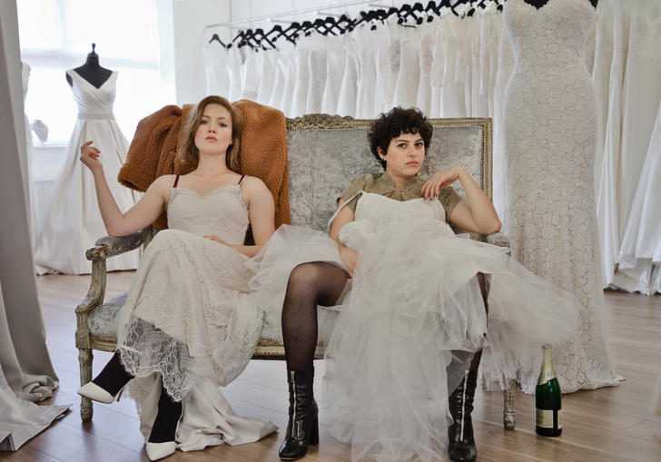 Holliday Granger and Alia Shawkat sitting in a wedding dress shop wearing wedding dresses pulled up to their knees