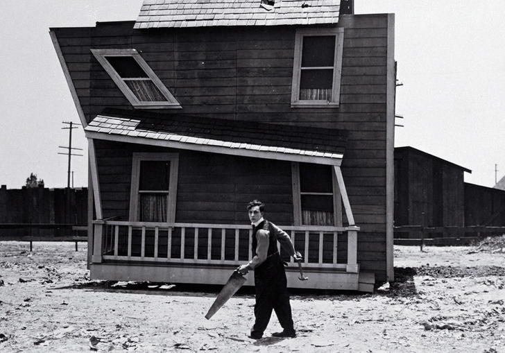 Buster Keaton stands with an saw outside a ramshackle, wonky wooden house