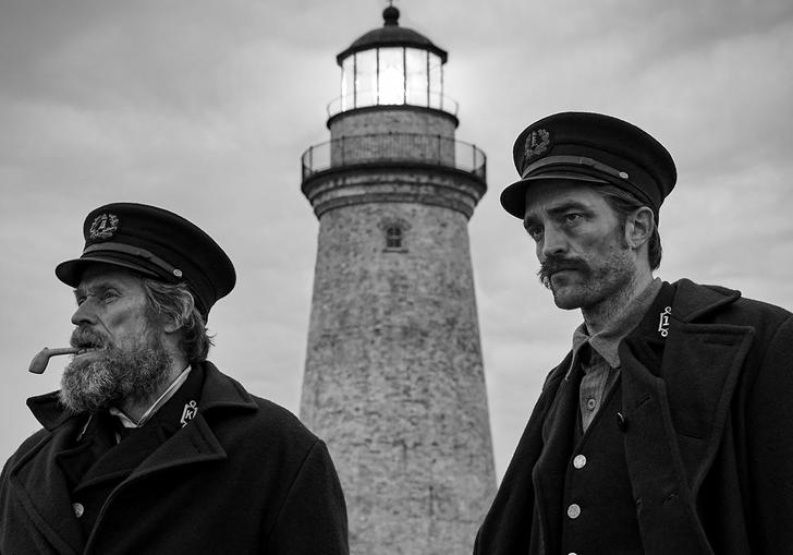 A black and white photo of Willem Dafoe and Robert Pattinson looking into the distance with a lighthouse behind them