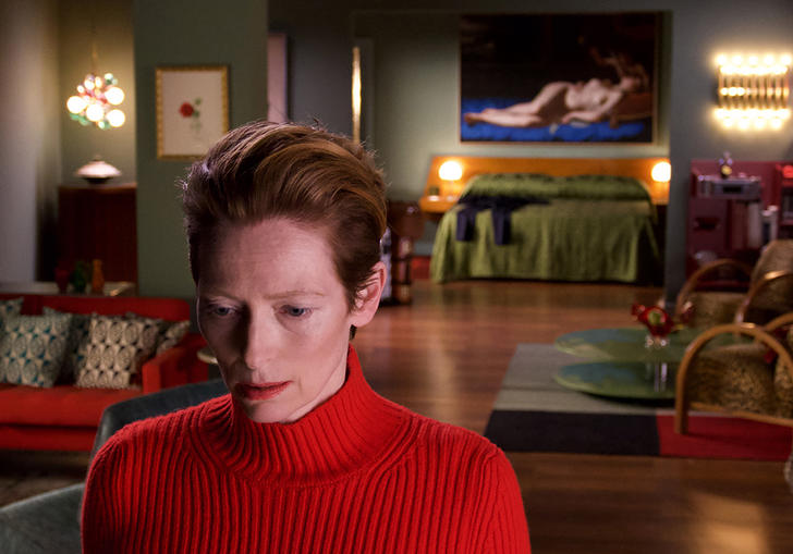 tilda swinton in a bright red top and swept back hair standing in an apartment