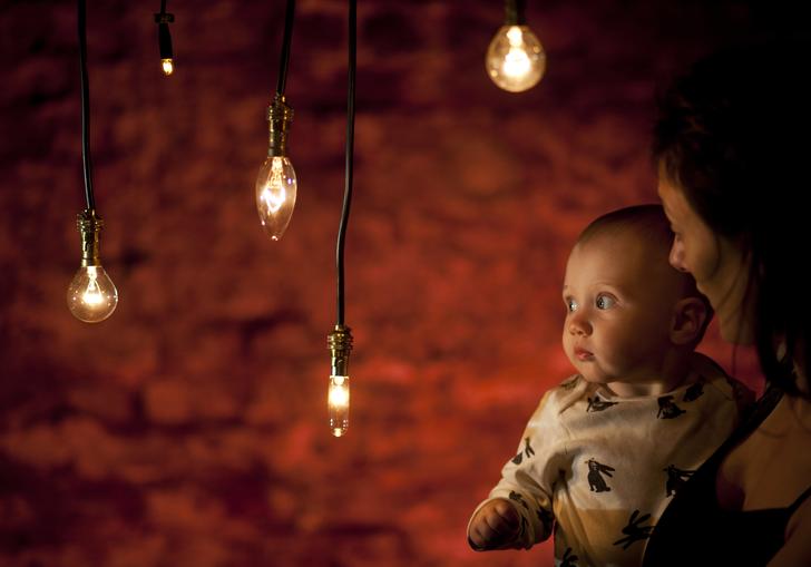 A baby in the arms of their mother look at hanging tungsten style light bulbs, bathed in a warm glow. Behind them is a red exposed brick wall. The mother is looking intently at the child.
