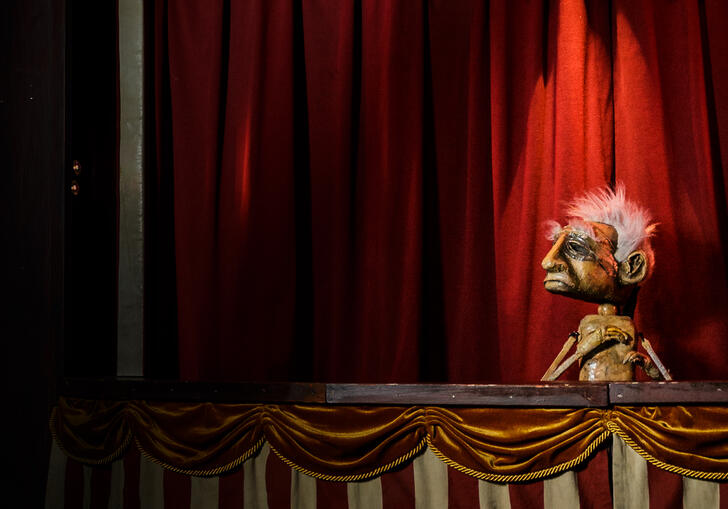 A rather sad looking puppet stands in a booth with a sign above their head that reads 'existential anguish'.