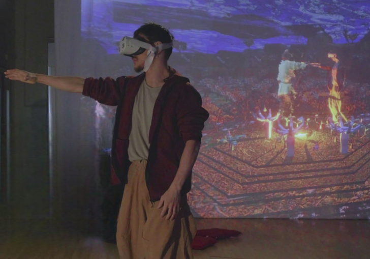 A person wearing a VR headset stands in front of a projector screen with their arm reaching towards something. On the projector screen the person appears in a 3D virtual world, reaching out to another person.