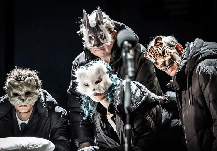 Four people wearing black coats and animal face masks gather together as if listening to or watching someone.