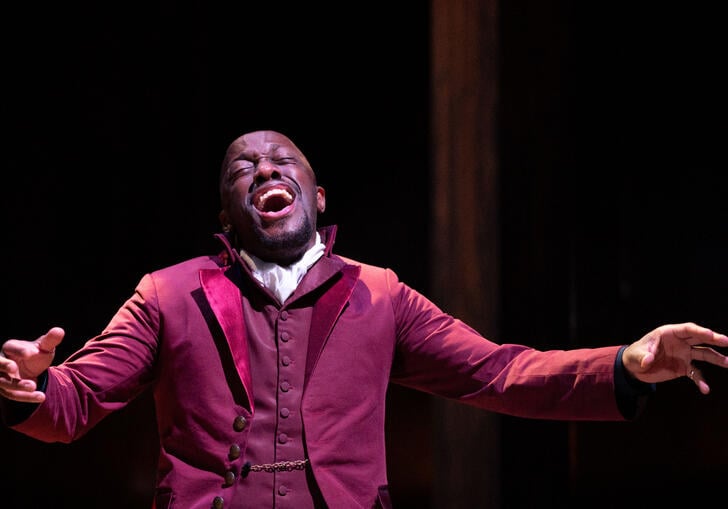Actor Giles Terera sings profoundly with his arms open while kneeling on stage in a red jacket