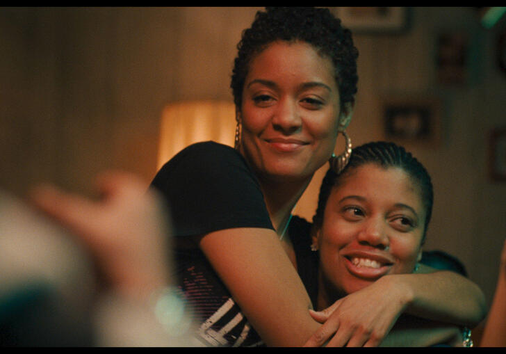 Two women cuddle on bed in a softly lit bedroom, smiling. 