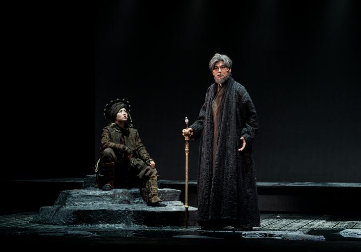 Lear stands with a wooden stick looking out at the audience while another person dressed in a dark green headdress sits on a stone beside him.