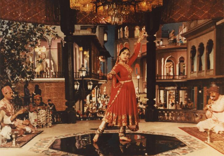 A woman in traditional Indian dress poses in a wonderfully decorated room, whilst men watch on.