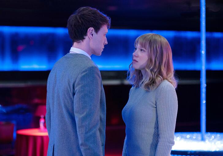 A smartly dressed man and a woman gaze at each other in a futuristic dark room.