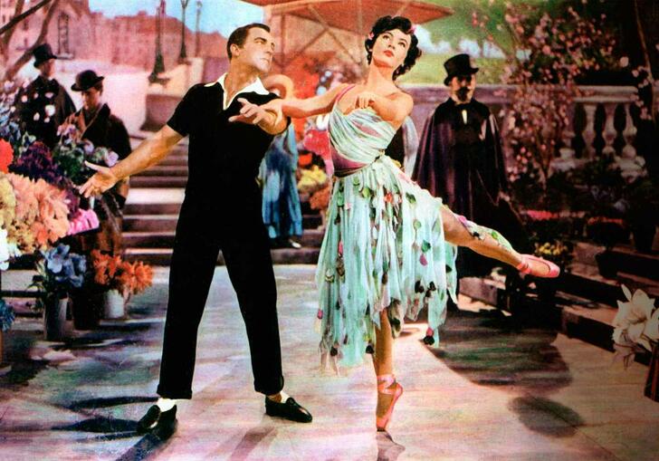 Two performers stand on stage holding hands, a woman wearing a blue dress takes a ballet stance, the man, in a black outfit watches her. 