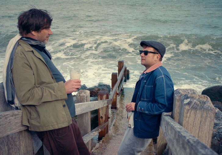 Two men stand on the stairs of a beach chatting, over a stormy sea.