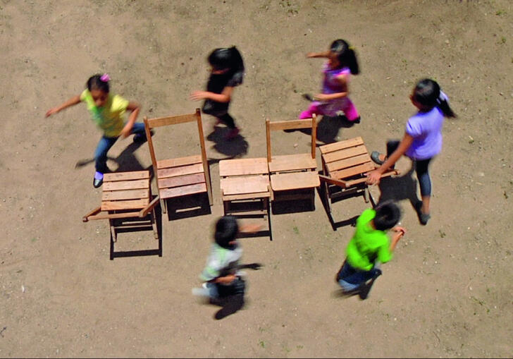 6 children running around 4 chairs, playing musical chairs