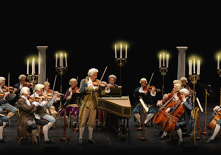 The orchestra performing on stage wearing period costume, with a row of tall candelabras behind them